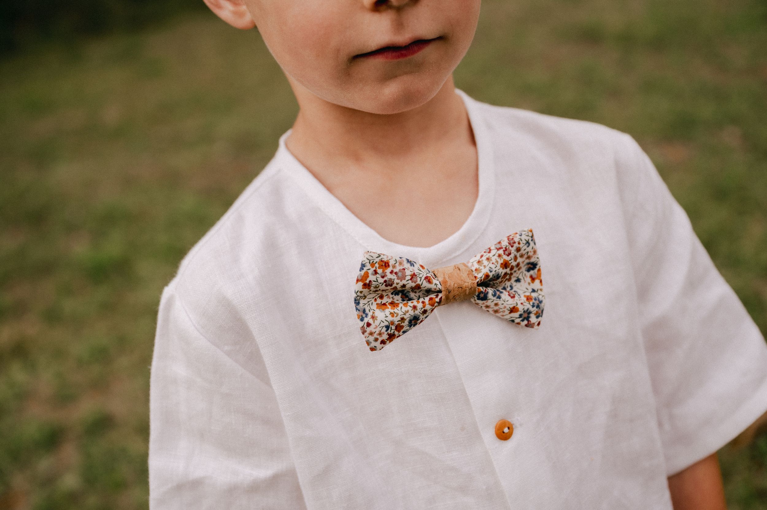 A child wearing a white shirt with a floral print bow tie standing in a grassy outdoor setting.