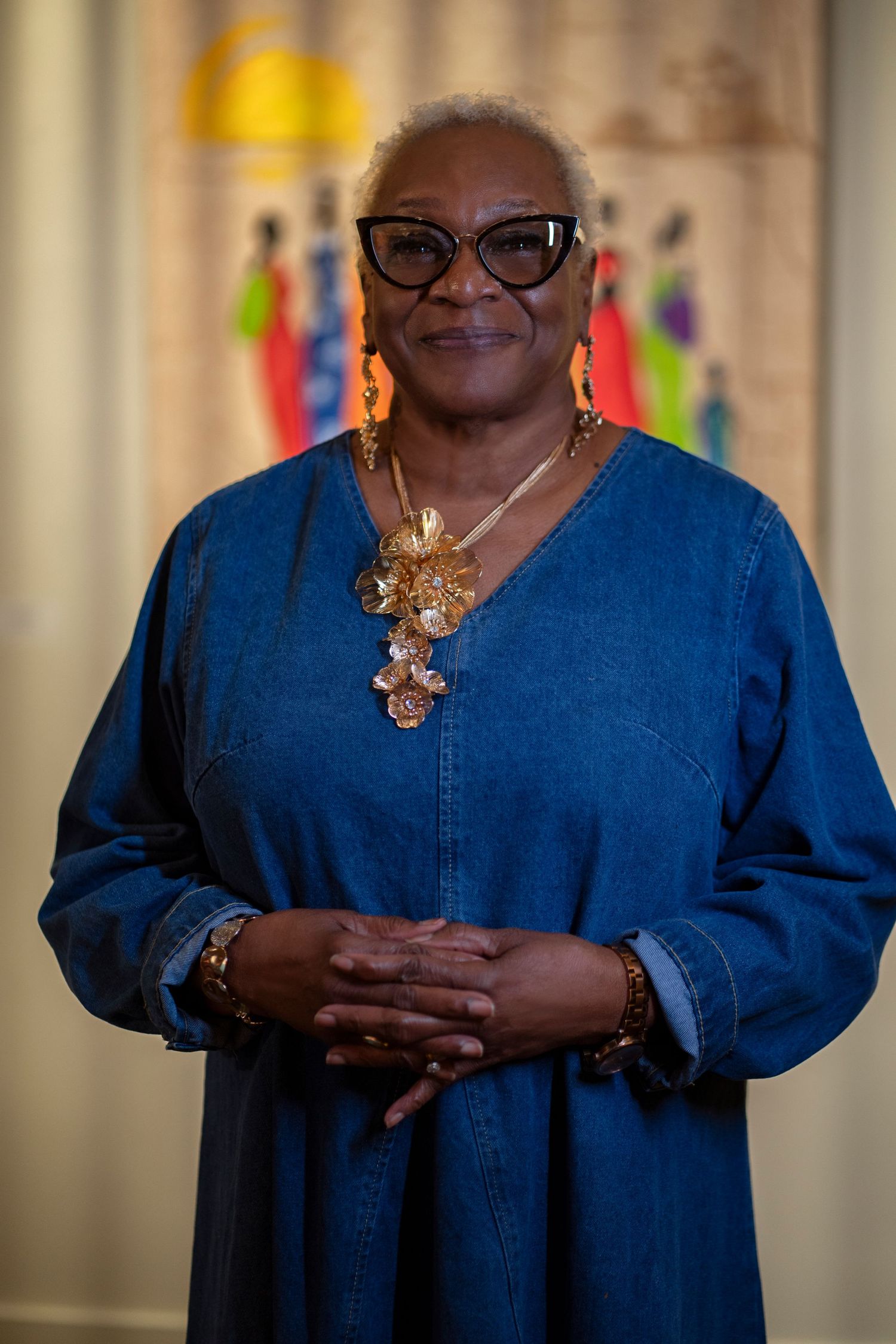 Person wearing blue denim dress and ornate gold necklace poses in front of patterned curtains.