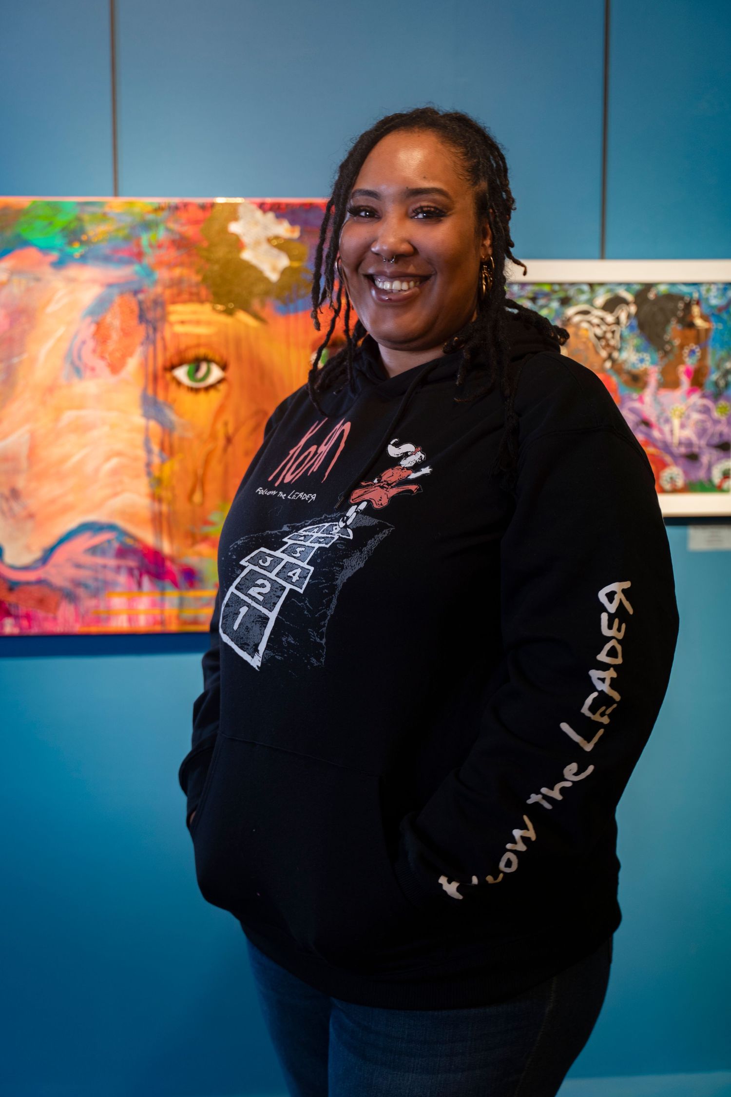 Artist stands smiling in black hoodie against blue wall with colorful artwork displayed in background.
