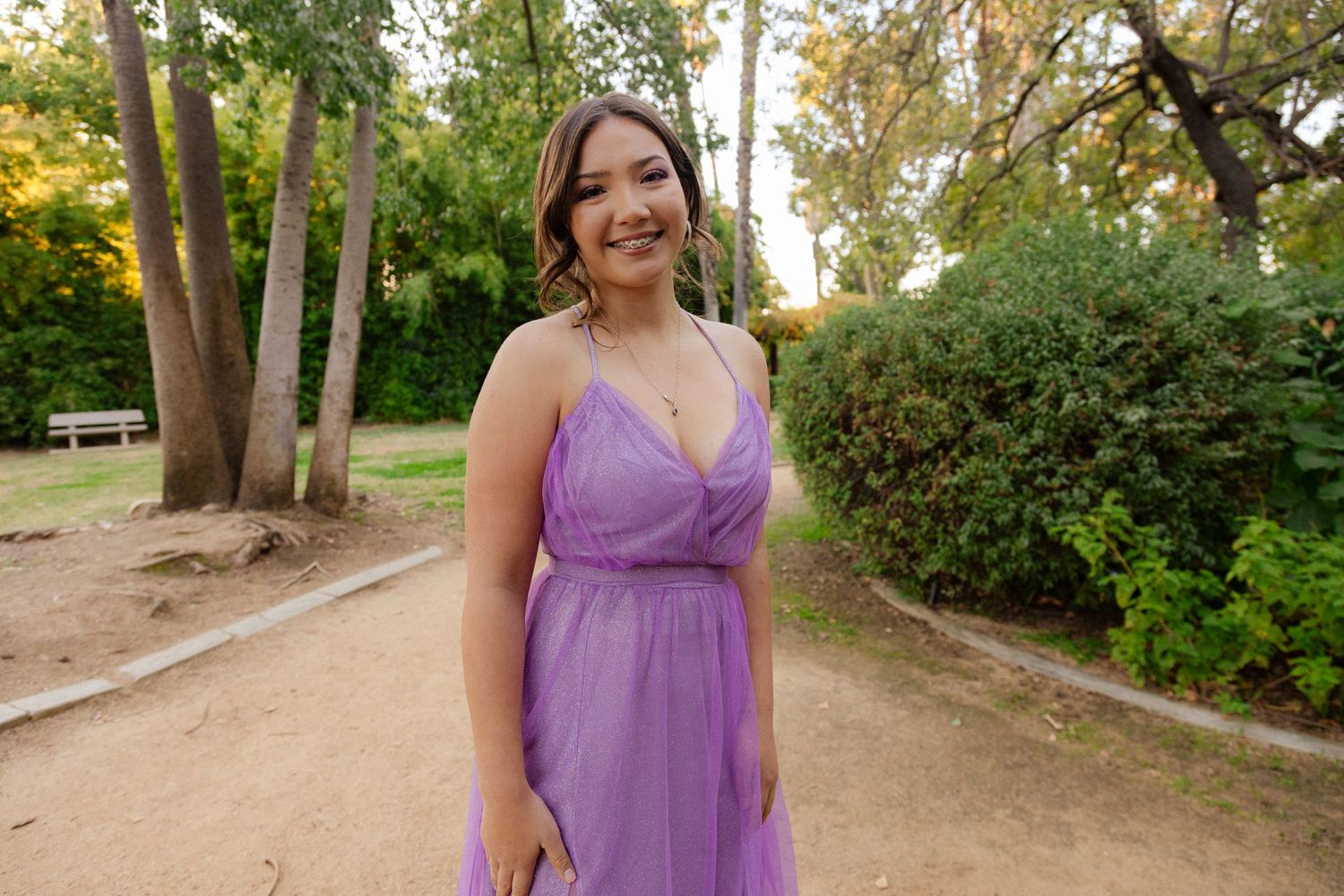 Woman in flowy light purple dress smiling on dirt path surrounded by trees in an outdoor park setting.