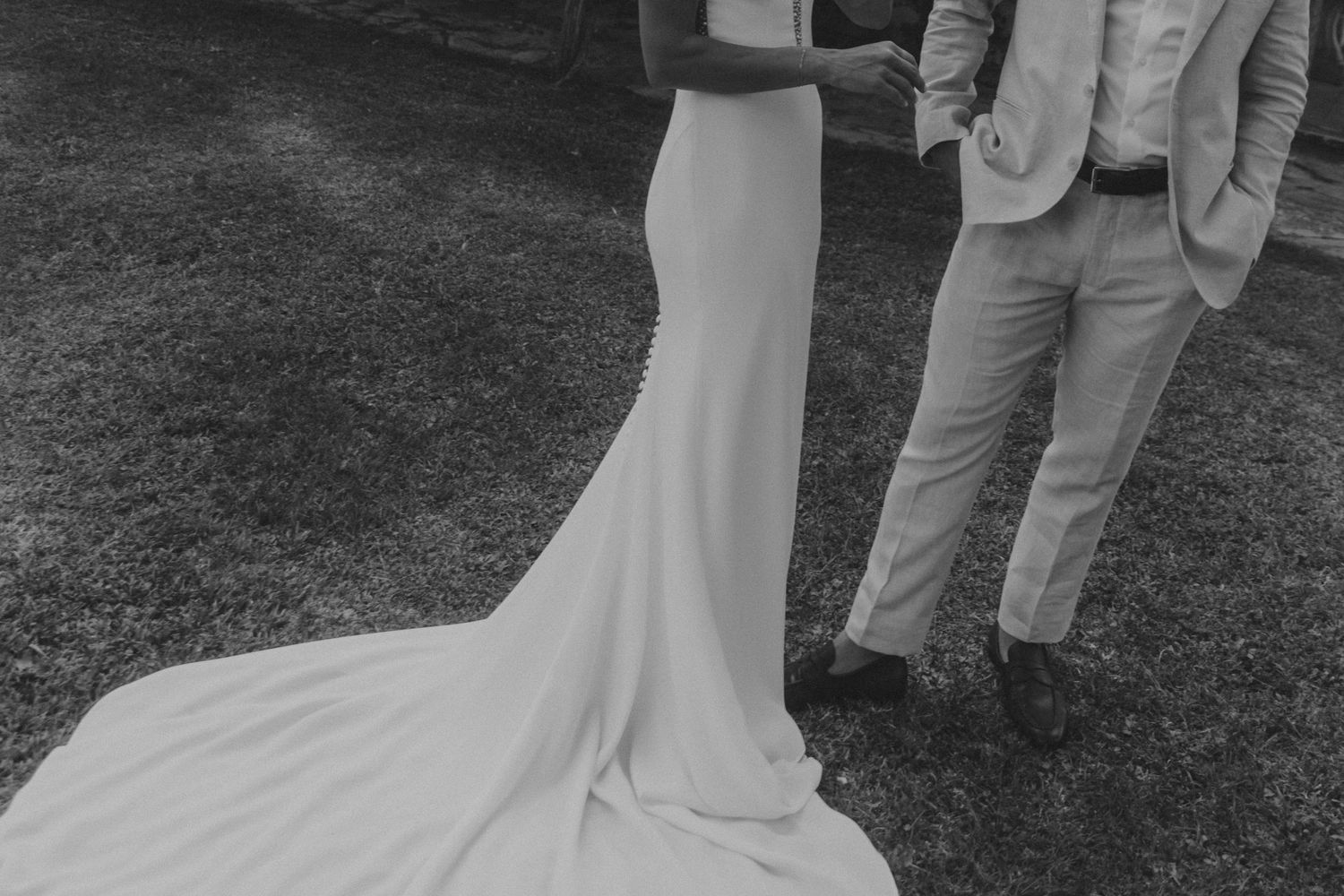 Black and white wedding photo showing the flowing train of a bride's dress next to a groom in a light colored suit.