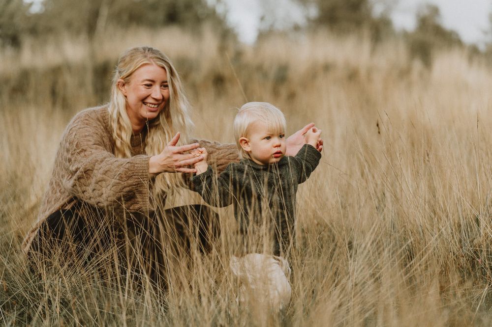 Mamma och barn står leker bland högt guld gräs under en familjefotografering i Jönköping. Mammans hår blåser i vinden.