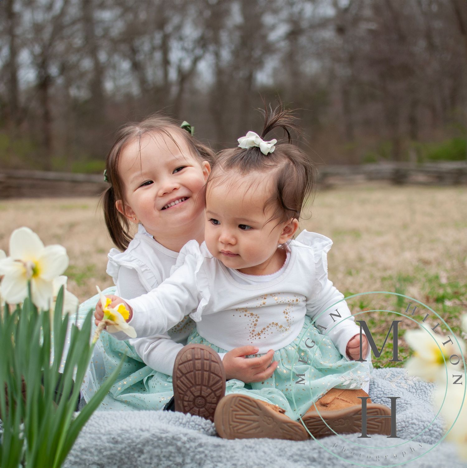 Photo of two young girls sitting on a blanket outdoors in spring with blooming daffodils, wearing matching white tops and smiling.