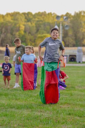 Children participate in an outdoor sack race on a grassy field during a sunny day with fall trees in the background.