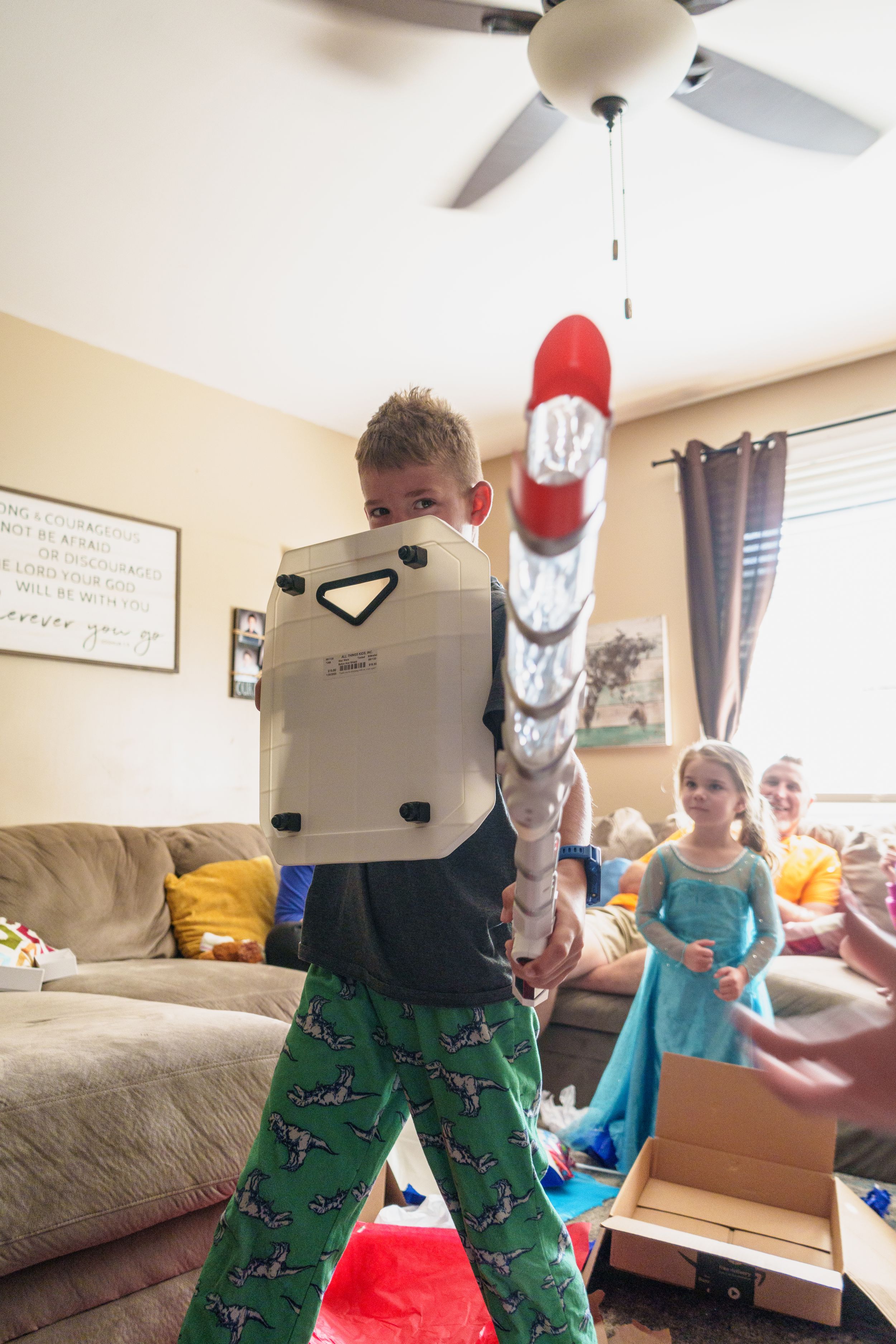 A child in Christmas-themed pajamas opens presents in a living room with a ceiling fan and holiday decorations.