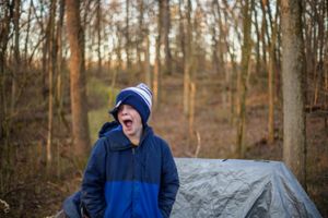 Someone in a blue winter fleece jacket and beanie standing in front of a gray tent in the woods during golden hour.