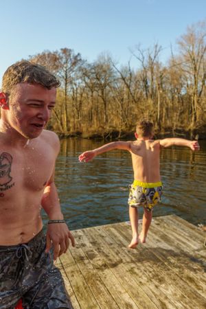 Two shirtless people stand on a wooden dock by a lake on a sunny autumn day preparing to jump into the water.