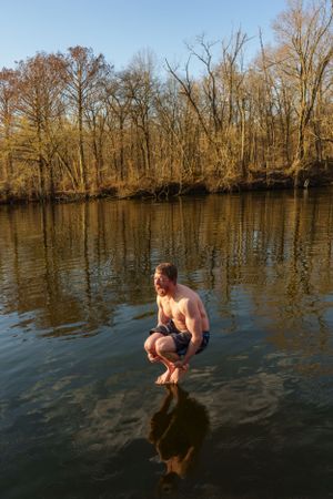 A person crouches on a partially submerged log in a tranquil river surrounded by bare winter trees at sunset.
