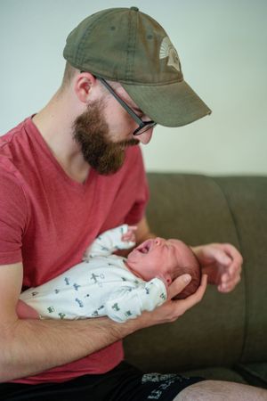 Parent wearing baseball cap and red t-shirt holds swaddled newborn baby while sitting on dark couch.