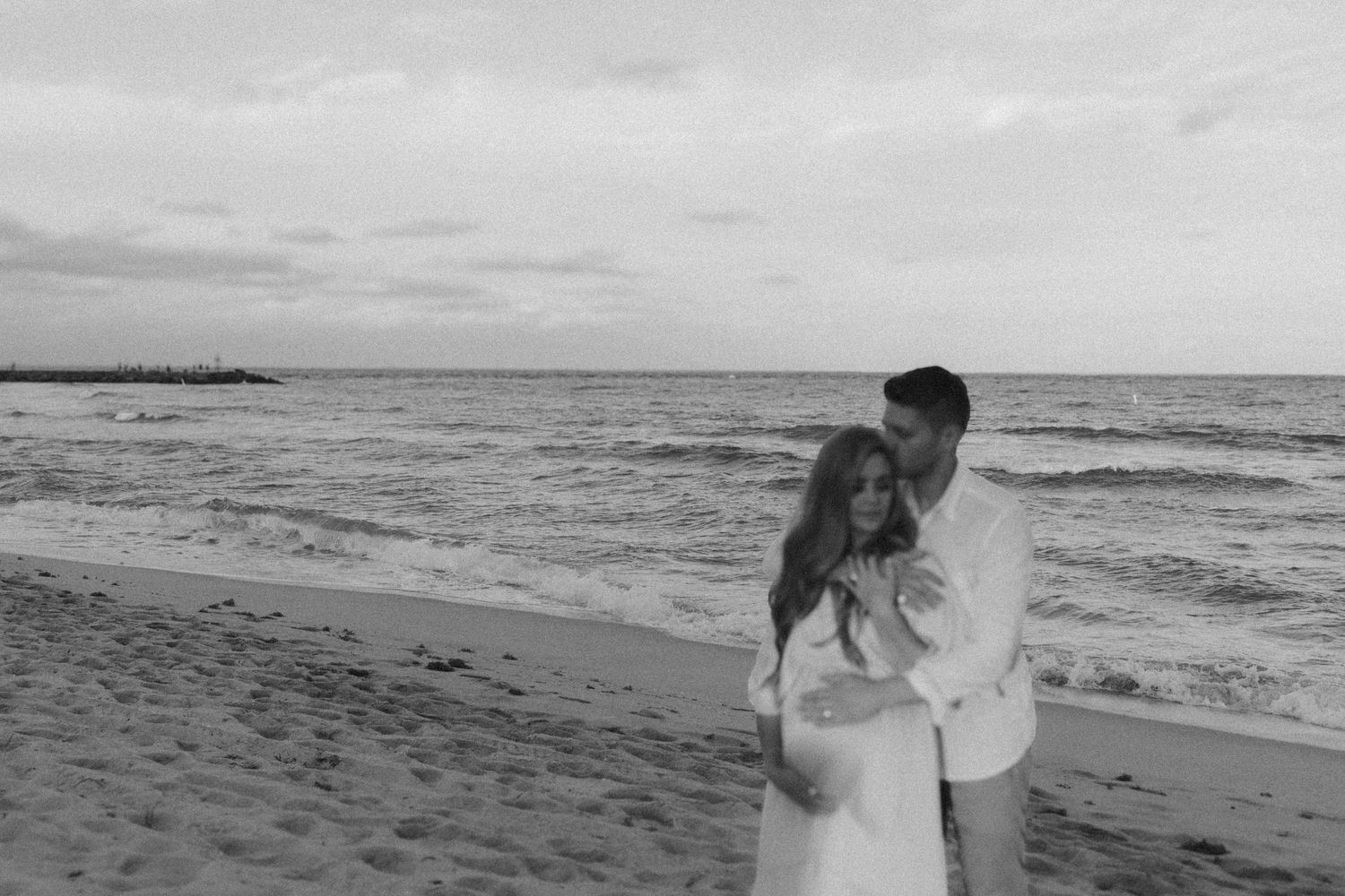 A couple embraces on a sandy beach during sunset with ocean waves in the background in black and white photography.