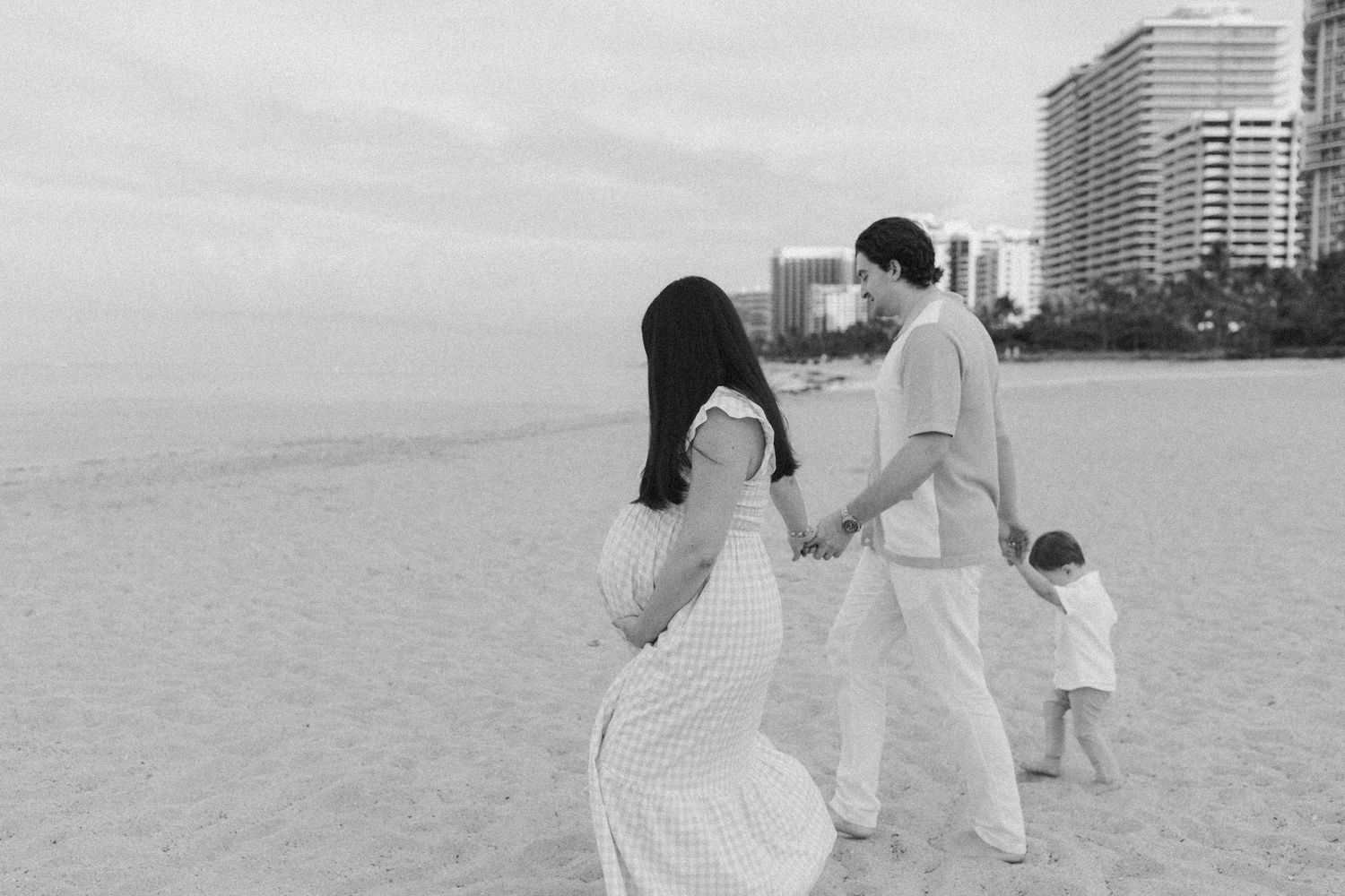 A couple embraces on a sandy beach during sunset with ocean waves in the background in black and white photography.