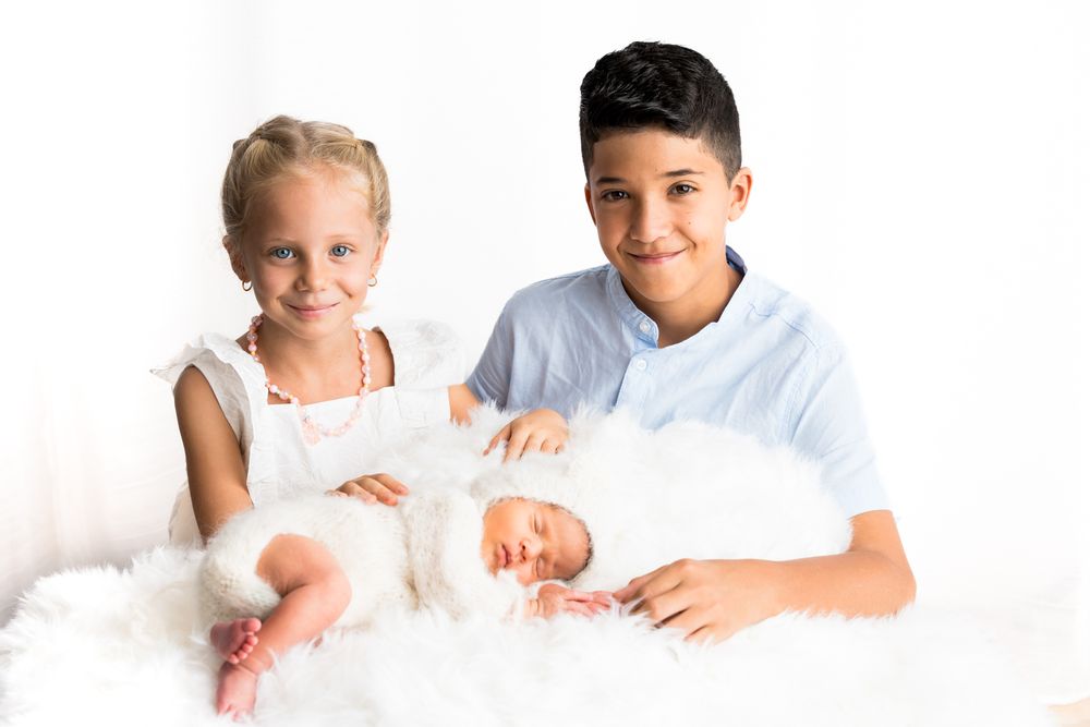 Séance Photo naissance Deux enfants tenant et regardant avec adoration un nouveau-né enveloppé dans une couverture blanche sur fond blanc.