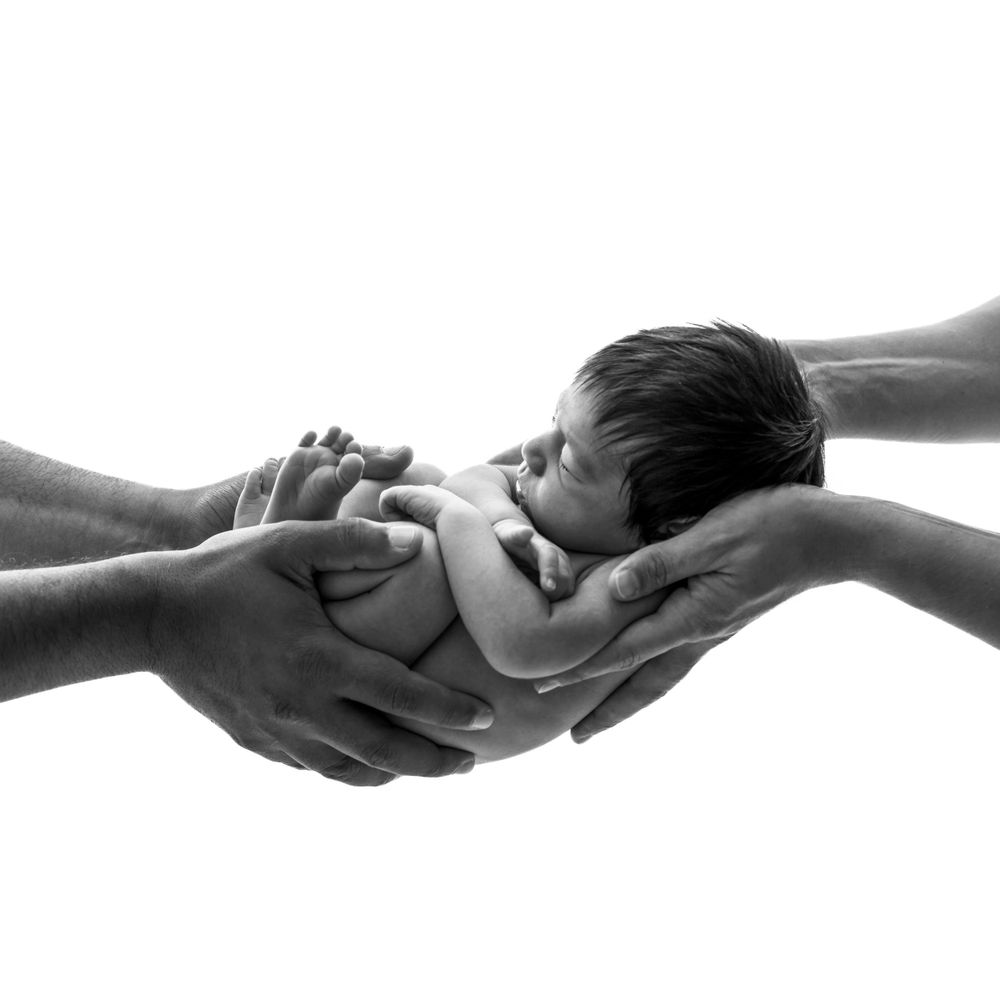 Séance Photo naissance  en noir et blanc d'un nouveau-né doucement bercé par plusieurs mains, symbolisant l'attention et le soutien.