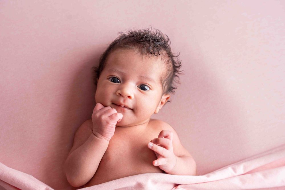 Séance Photo naissance Réunion : Doux bébé souriant allongé sur une couverture rose et suçant son pouce.