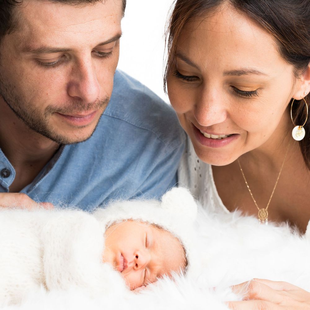 Séance photo naissance en studio pro :Moment tendre où les parents regardent avec amour leur nouveau-né endormi dans une couverture blanche.