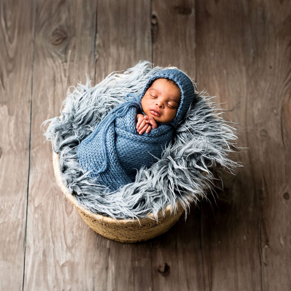 Photo naissance Newborn wrapped in cozy blue knit blanket sleeping peacefully in basket with fluffy gray fur lining on wooden floor.