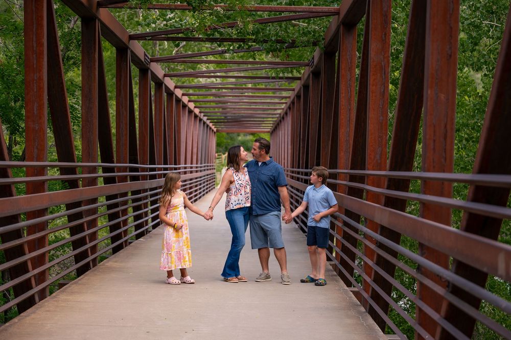 Family walking together across a rustic metal bridge surrounded by lush green trees on a sunny day.