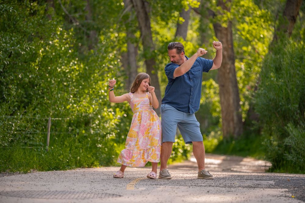 Two people dance joyfully together on a sunlit path through a green wooded area on a summer day.