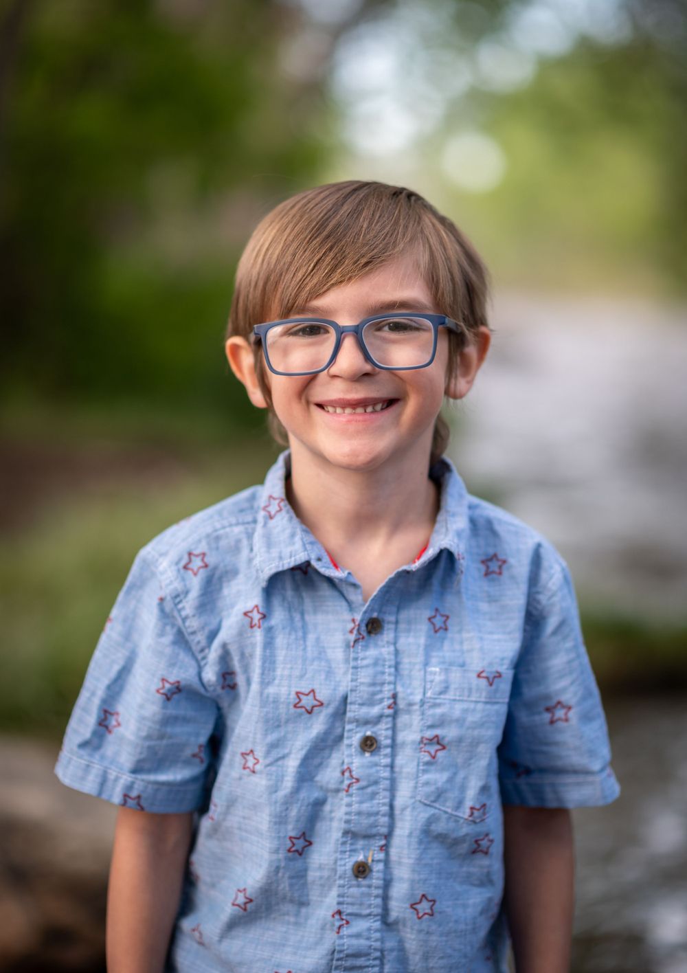A young person wearing blue rimmed glasses and a light blue patterned button-down shirt smiles outdoors in natural lighting.