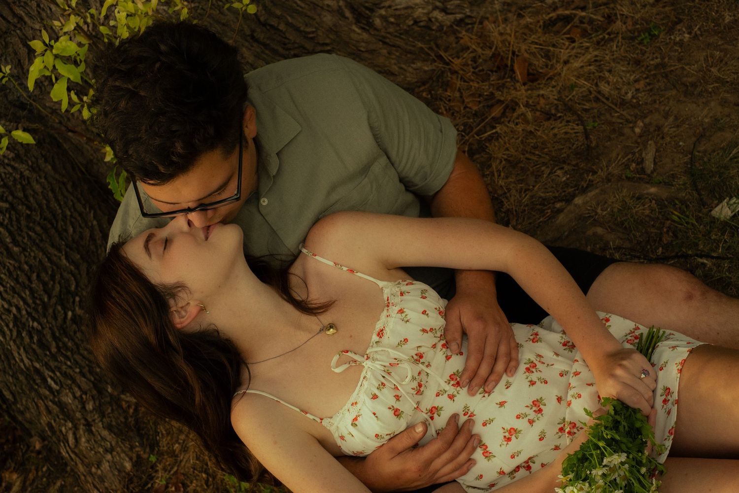 A romantic couple shares an intimate moment lying on the ground in a forest setting, bathed in soft natural light.