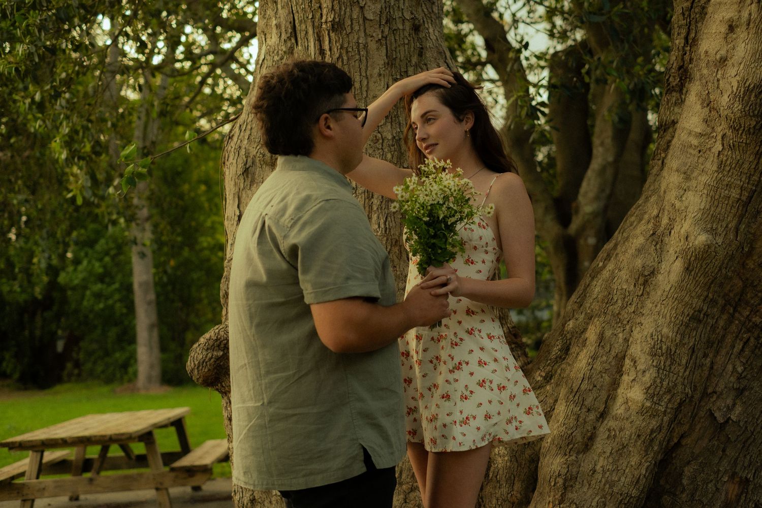 A couple shares a romantic moment beneath a large tree as one holds a bouquet of wildflowers.