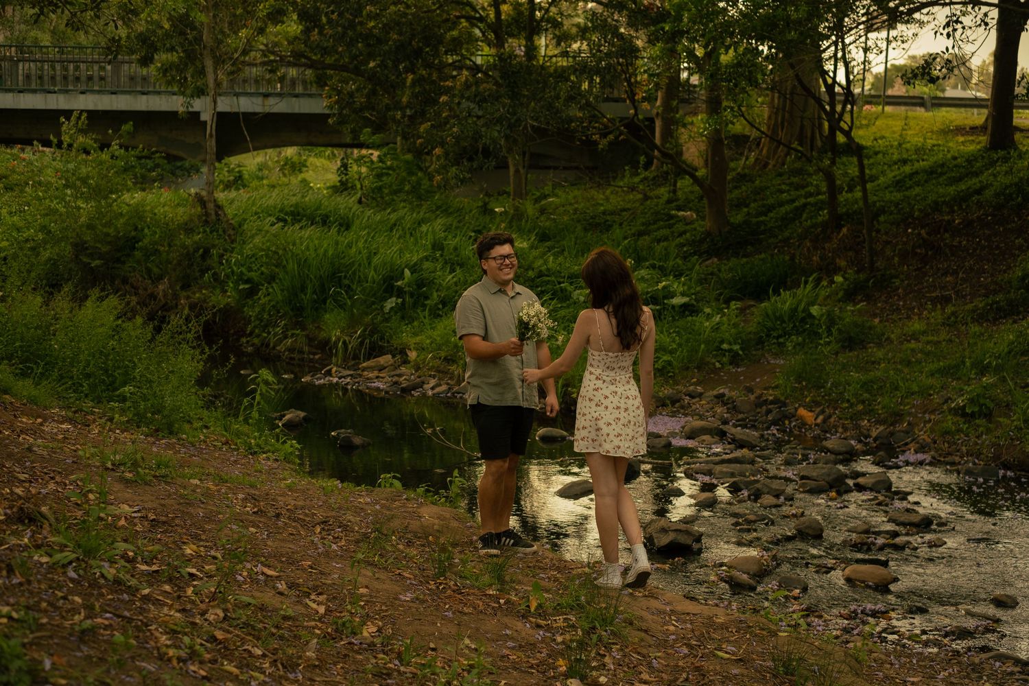 A couple stands together near a small stream in a lush green outdoor setting during golden hour.