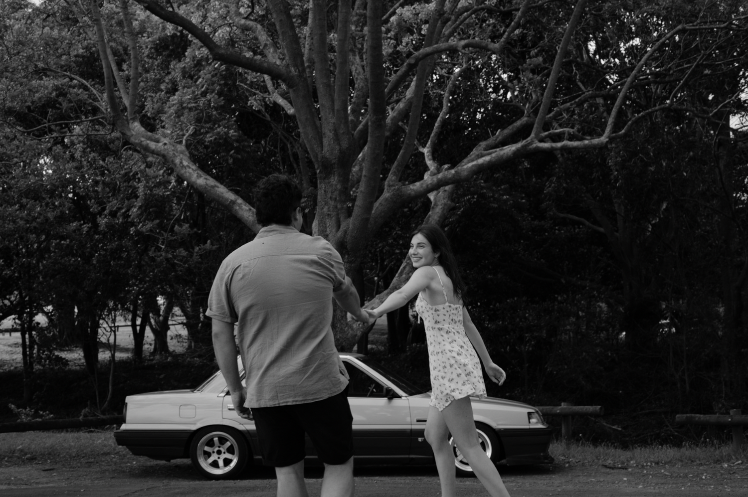 Two silhouettes stand near a car in a park setting with trees in black and white photography.
