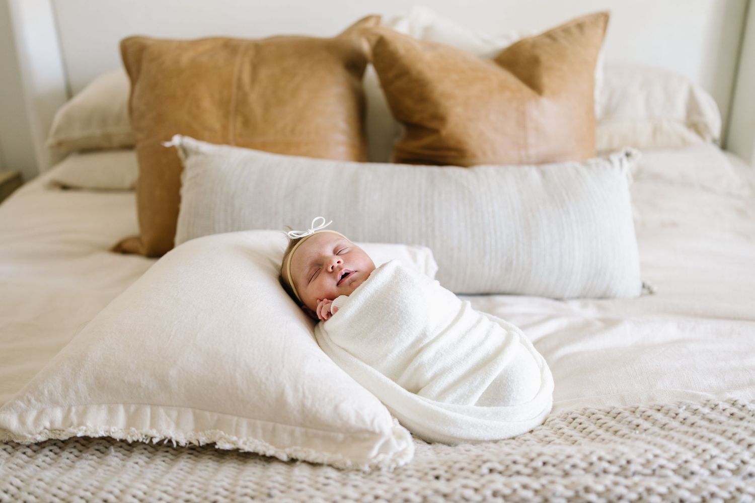 Newborn baby swaddled in white wrap lying on bed with tan pillows.