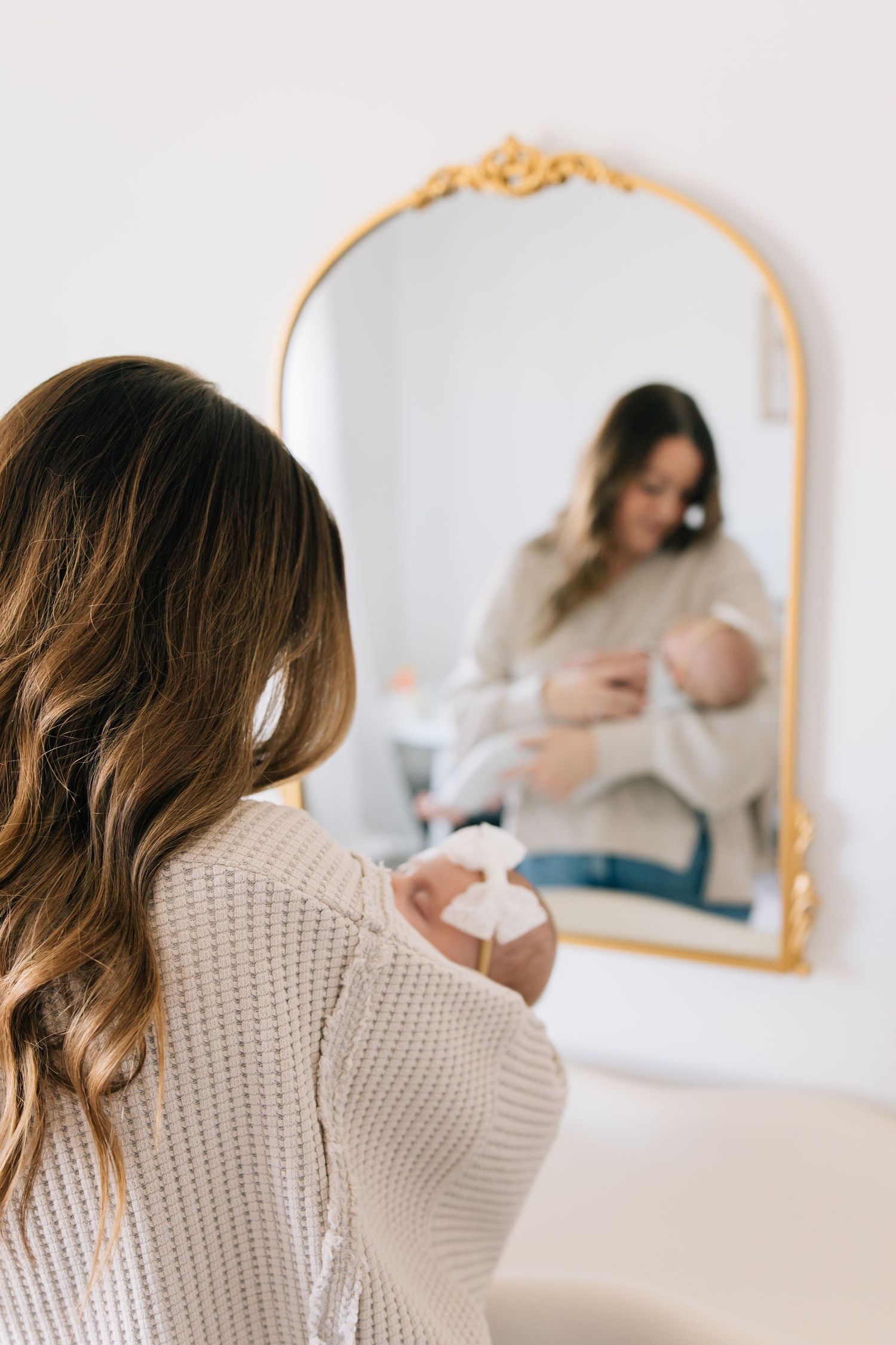 Woman in knit sweater breastfeeding baby reflected in ornate gold mirror against white wall.