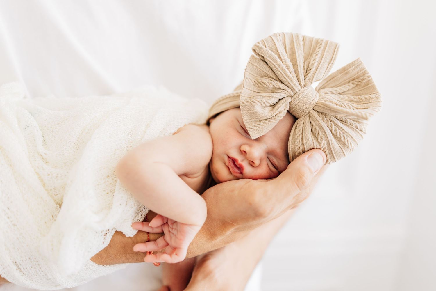 Sleeping baby wearing a large beige bow wrapped in white fabric.