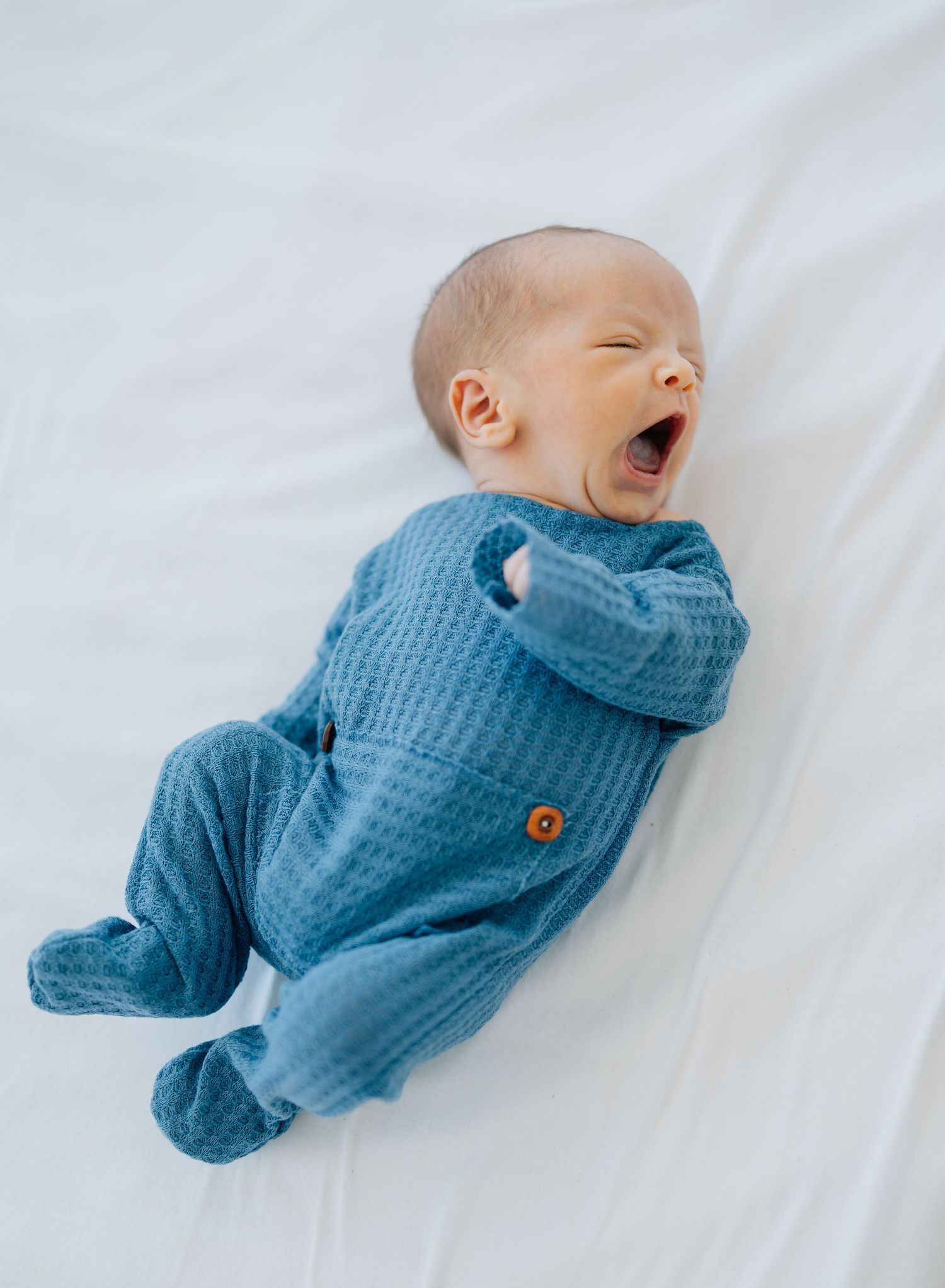 Baby in blue knit sleeper yawning while lying on white bedding.