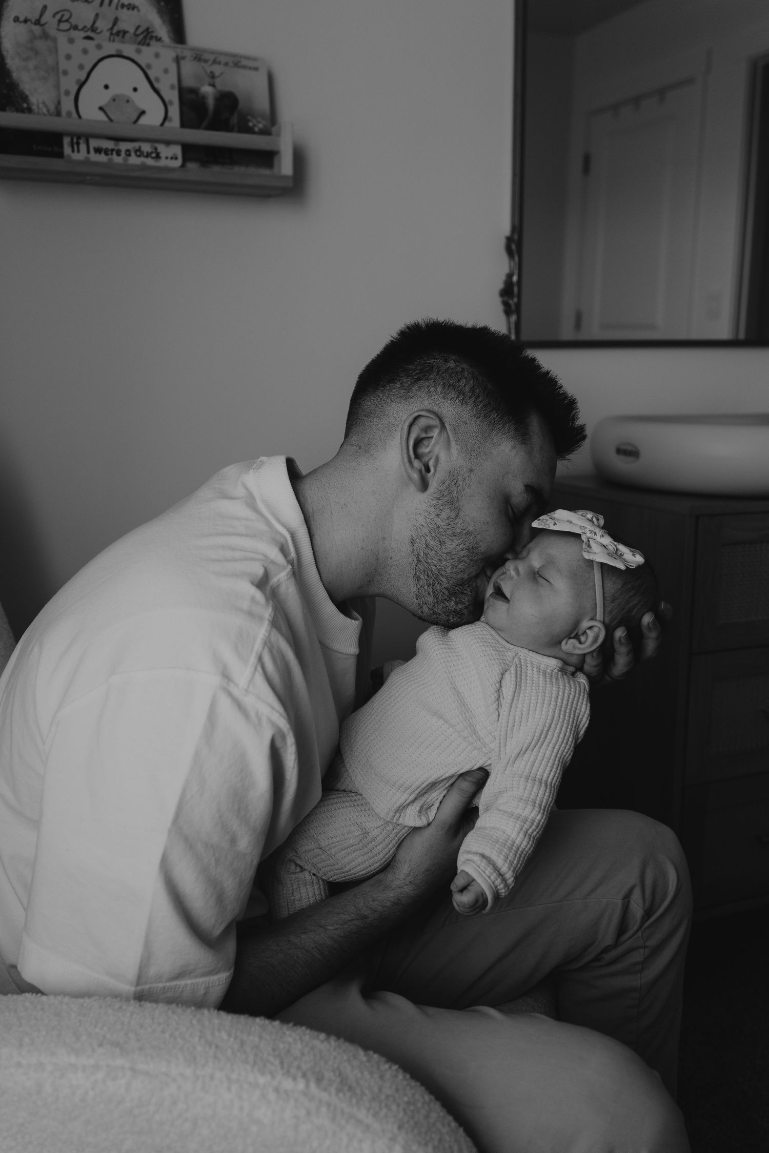 Tender black and white moment of parent kissing newborn baby in nursery setting.