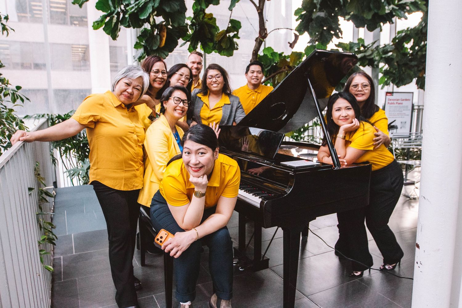 Group of women in yellow shirts posing around a grand piano indoors, surrounded by lush green plants.