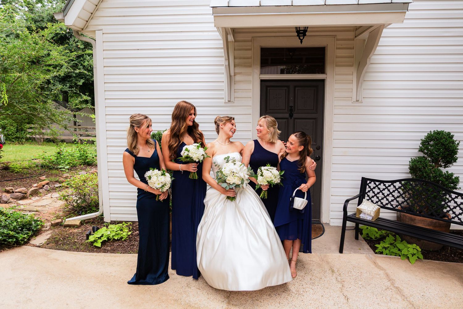 Bride in white gown with four bridesmaids in navy blue dresses holding white bouquets outside a white chapel.