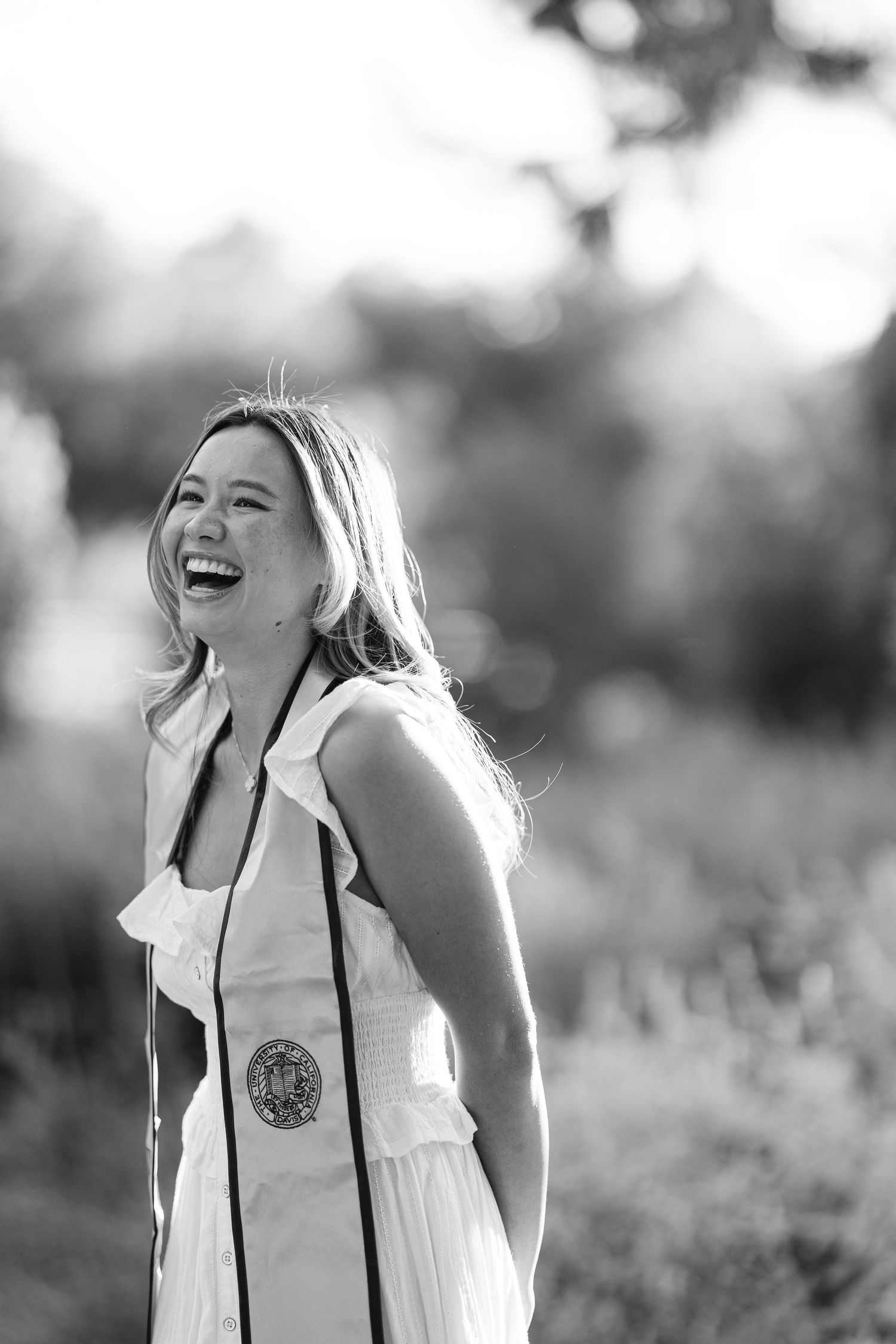 Black and white photo of a laughing graduate in a white dress with a medallion sash outdoors.