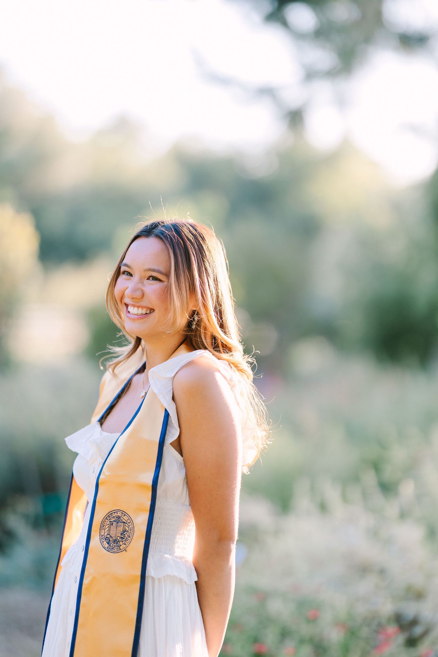 Smiling young woman in white dress wearing gold graduation stole poses outdoors in sunlit garden.