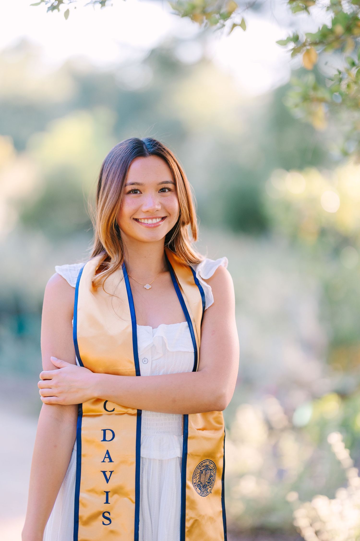 Smiling young woman in white dress wearing UC Davis graduation stole posing outdoors.