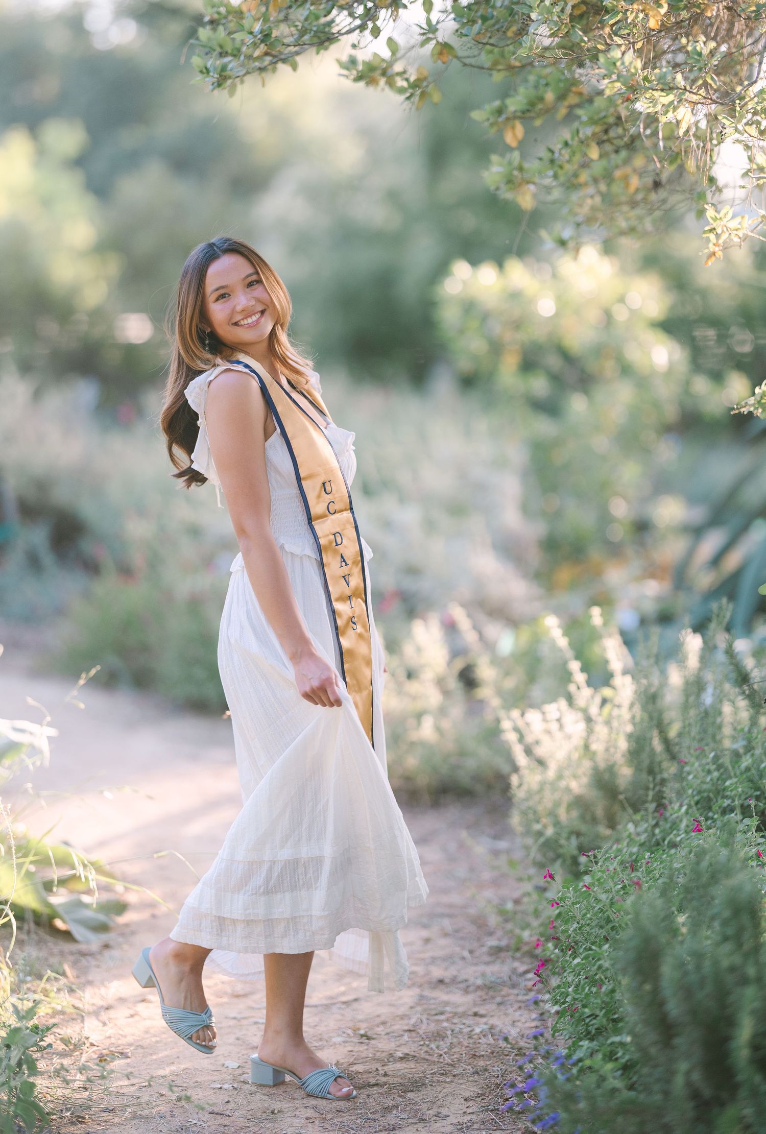Smiling graduate in white dress and gold graduation sash poses on a sunlit garden path surrounded by lush greenery.