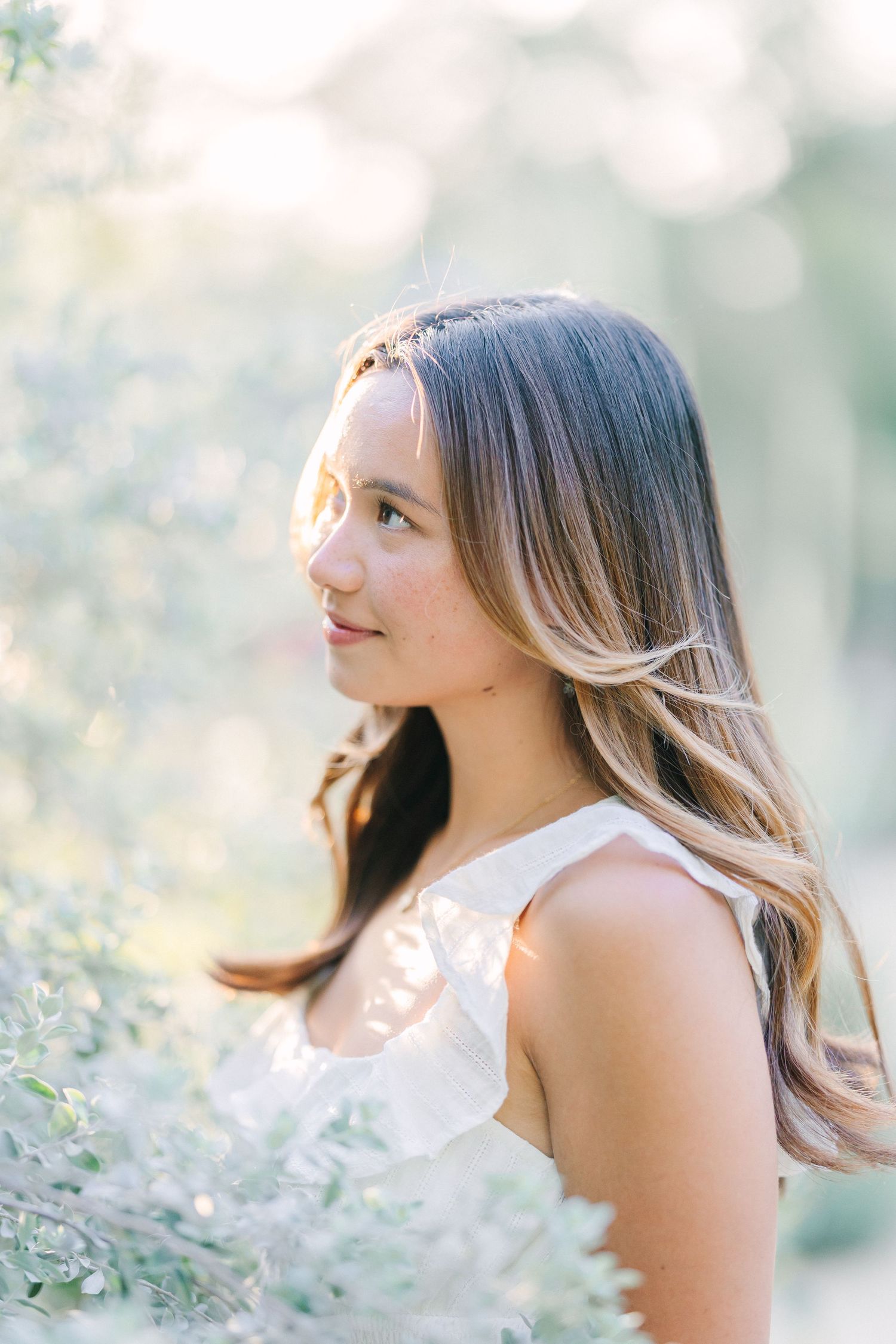 Young woman with long highlighted hair wearing a white dress, posed outdoors among soft green foliage in golden light.