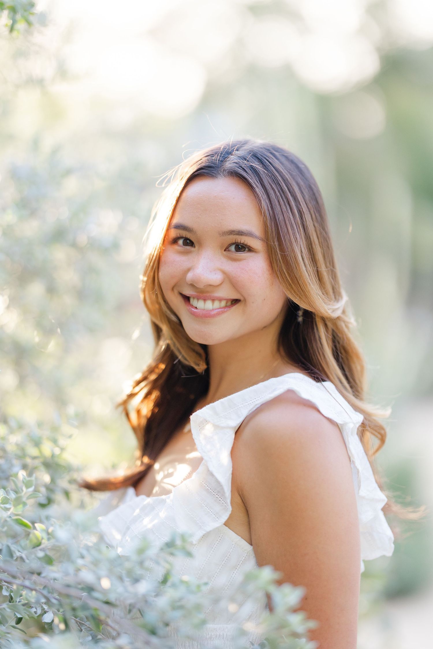 Young woman with long brown hair smiling outdoors in white ruffled dress, surrounded by soft green foliage in natural light.