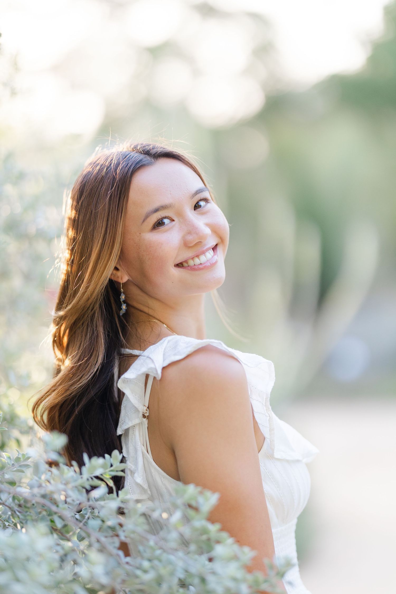 Smiling young woman senior portrait in white dress outdoors with soft bokeh background and warm natural light.