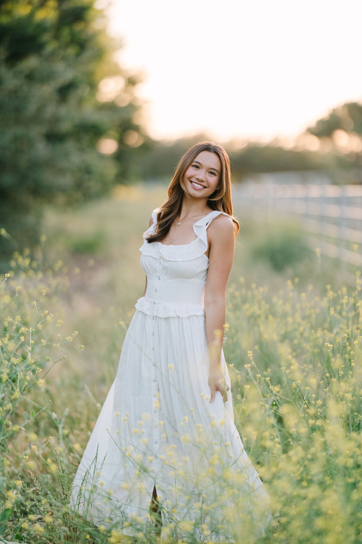 Happy young woman in white ruffled dress smiles in golden hour sunlight among wildflowers.