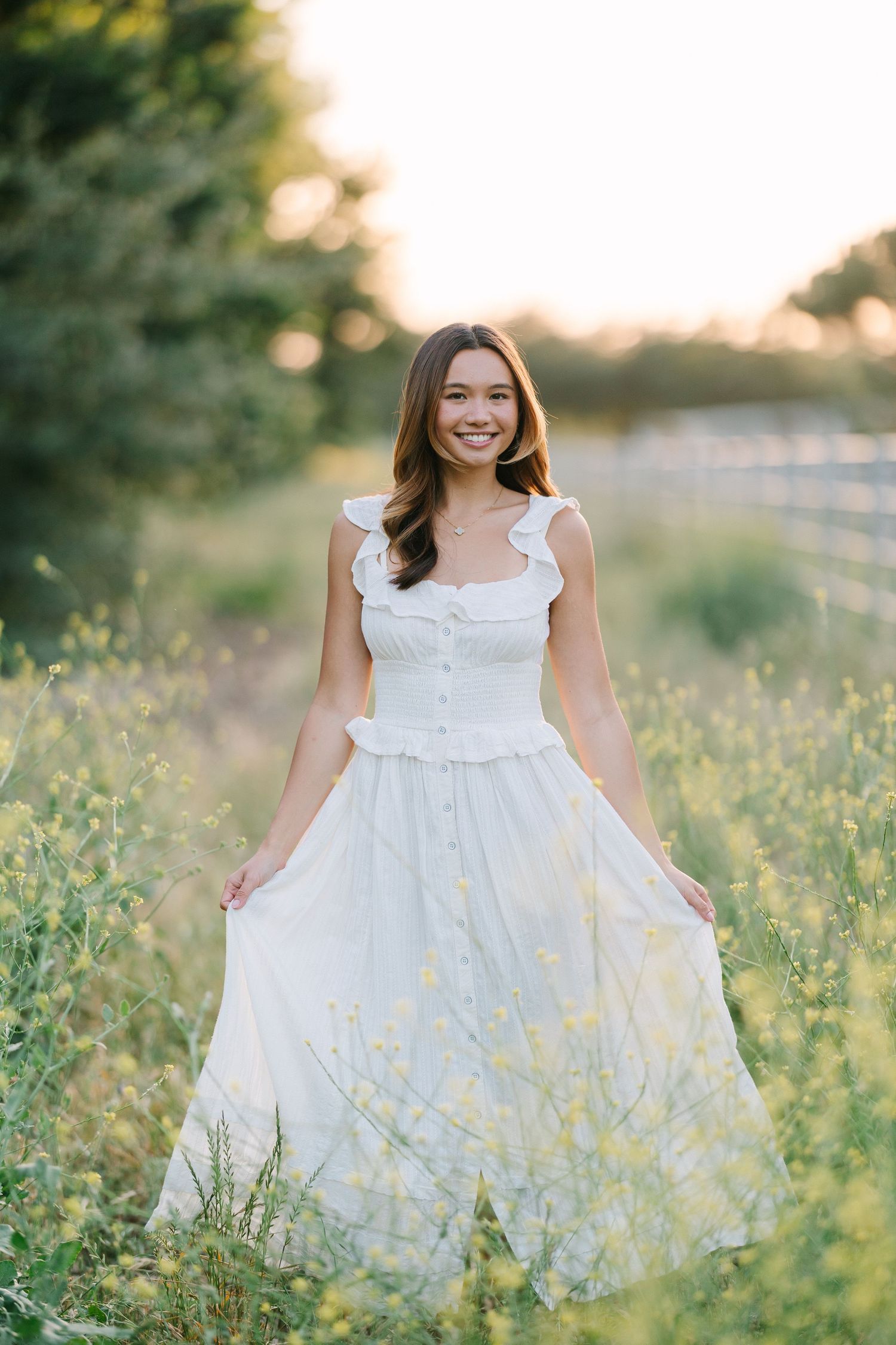 Young woman in white ruffle dress smiling in a sunny field of wildflowers at golden hour.