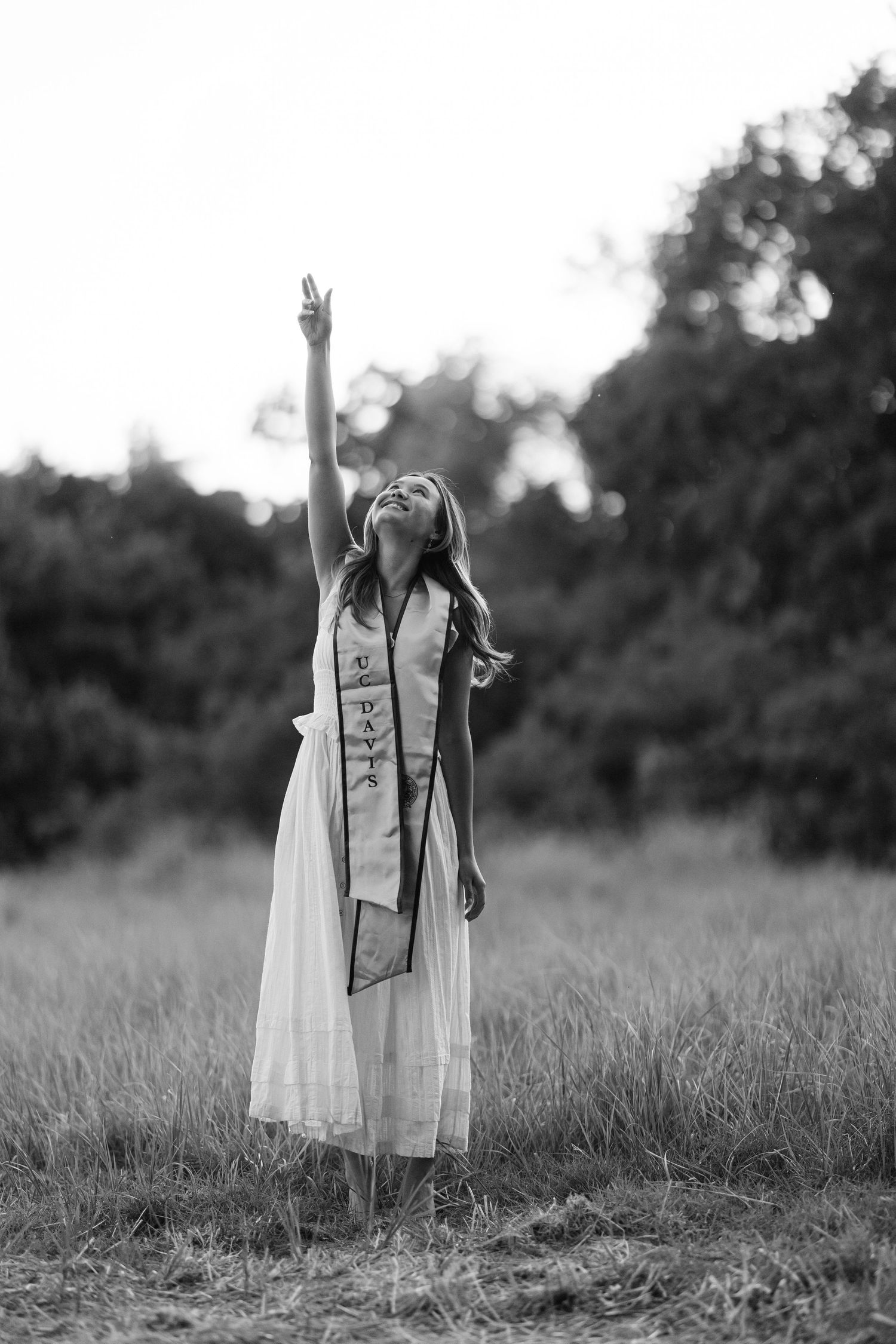Young woman in white dress and graduation sash reaches toward sky in black and white outdoor photo.