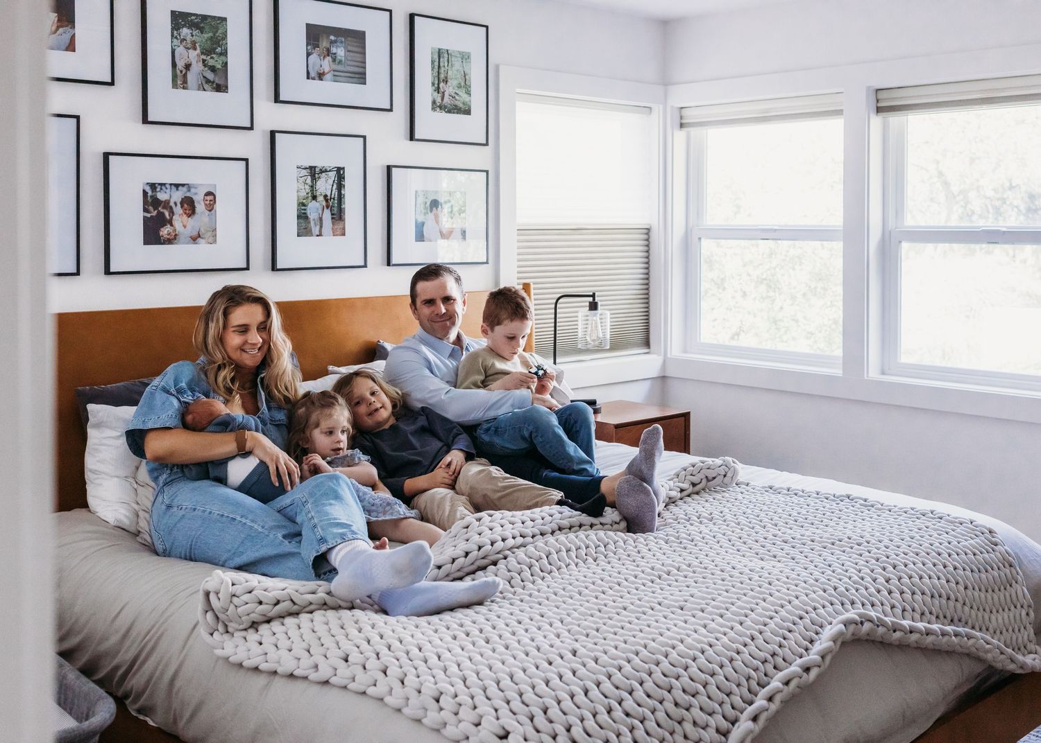 Happy family of five lounging together on a bed with chunky knit blanket, gallery wall of photos behind them.