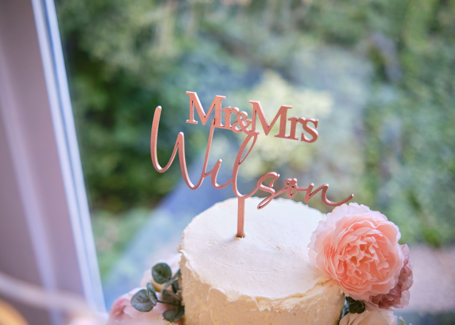 Rose gold Mr & Mrs Wilson cake topper on rustic wedding cake with pink floral decorations.