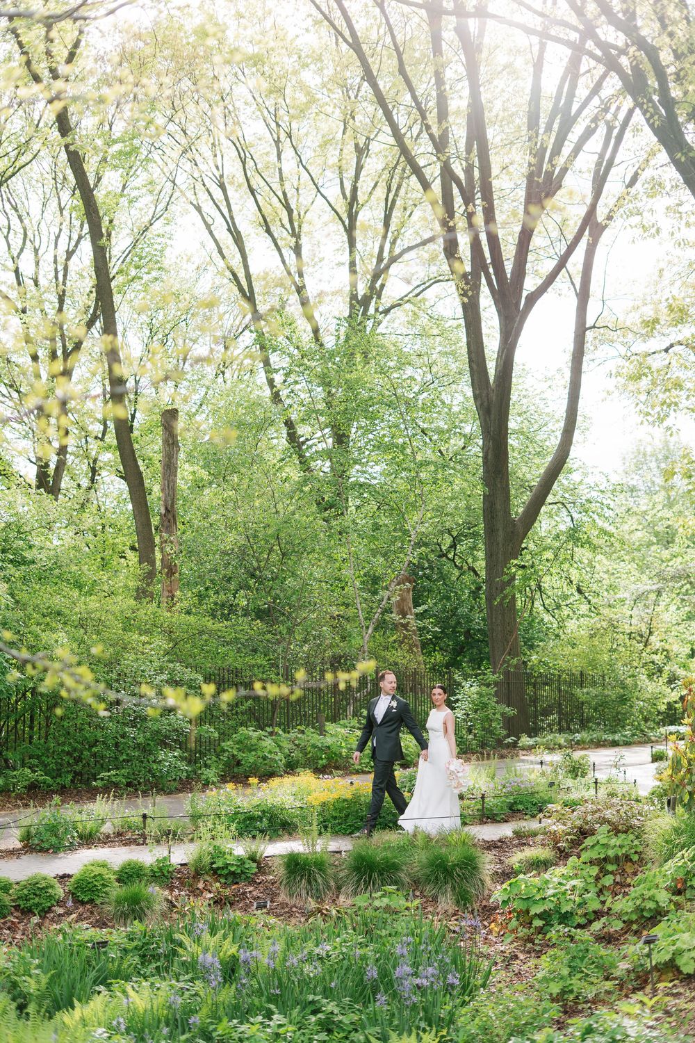 Bride and groom walk hand in hand through a lush green garden on their wedding day.