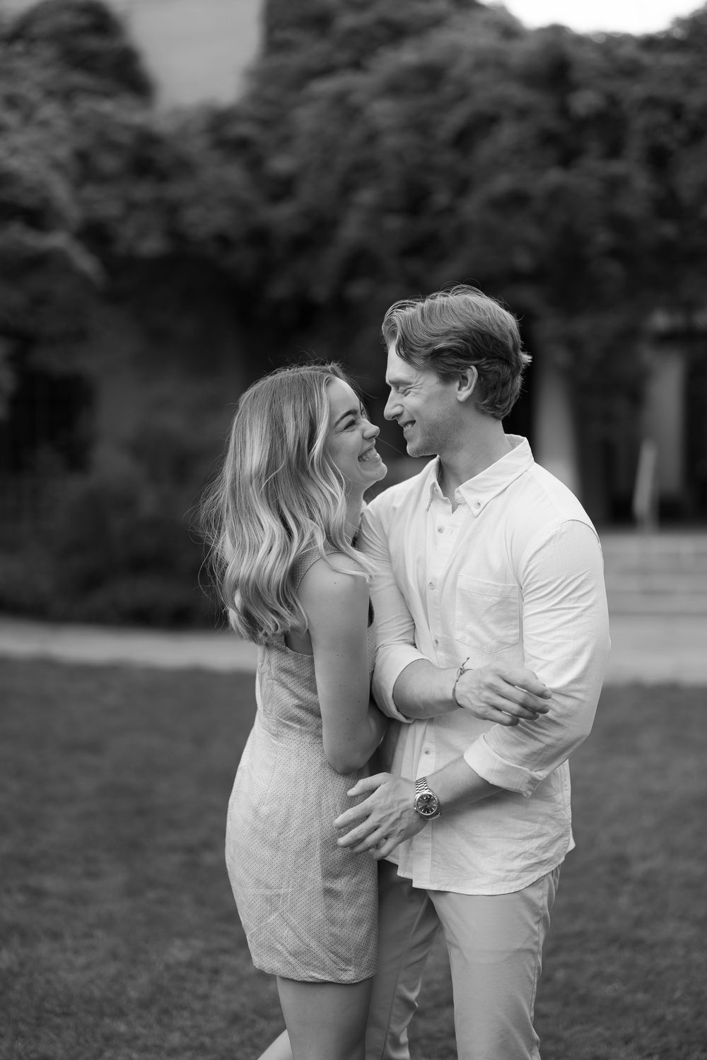 Black and white photo of a smiling couple embracing outdoors on a lawn with lush green trees in the background.