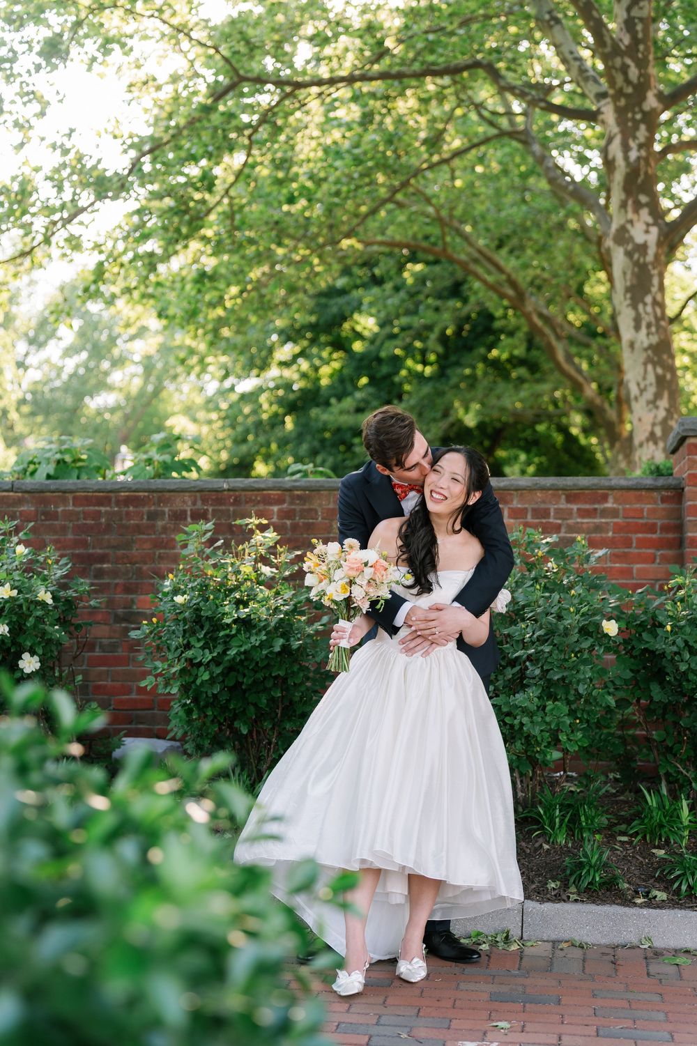 Bride and groom embrace in lush garden, she holds floral bouquet in white dress as he kisses her cheek.