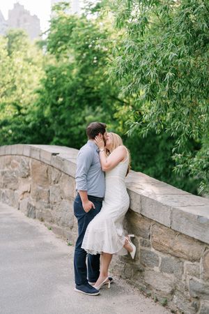 Couple sharing a romantic kiss against a stone bridge wall surrounded by lush green trees in Central Park.